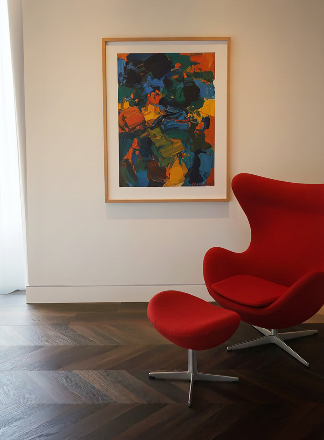 Red chair and ottoman on a dark walnut chevron floor, with a colorful abstract painting on a white wall.
