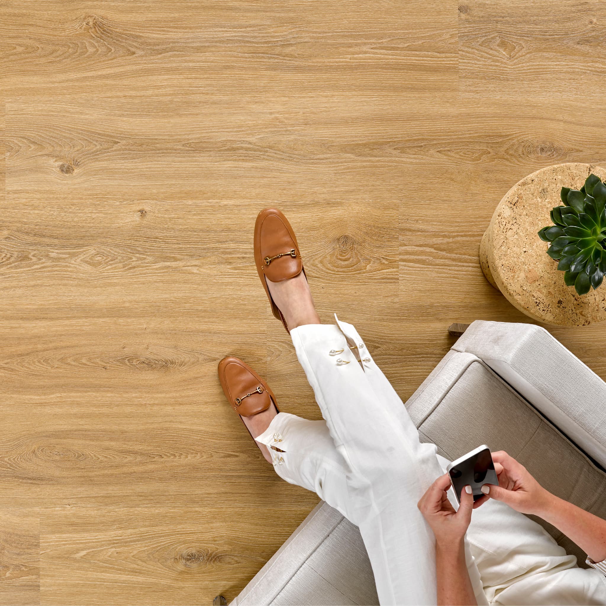 Person sitting on a couch with brown shoes and white pants, holding a phone on an Allure LVP floor.