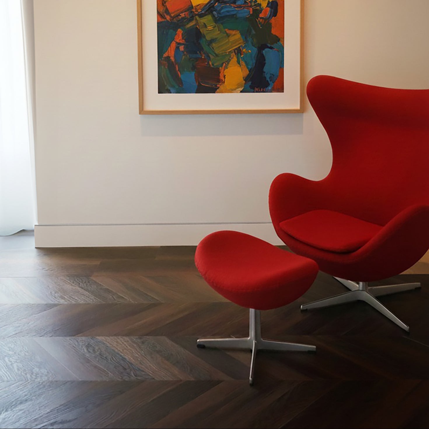 Red chair and ottoman on a dark walnut chevron floor, with a colorful abstract painting on a white wall.
