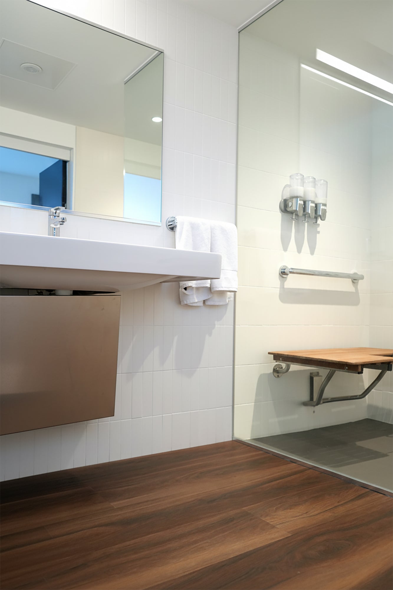 Modern commercial bathroom with Allure reddish wood LVP floor, mirror, towel rack and shower stall.